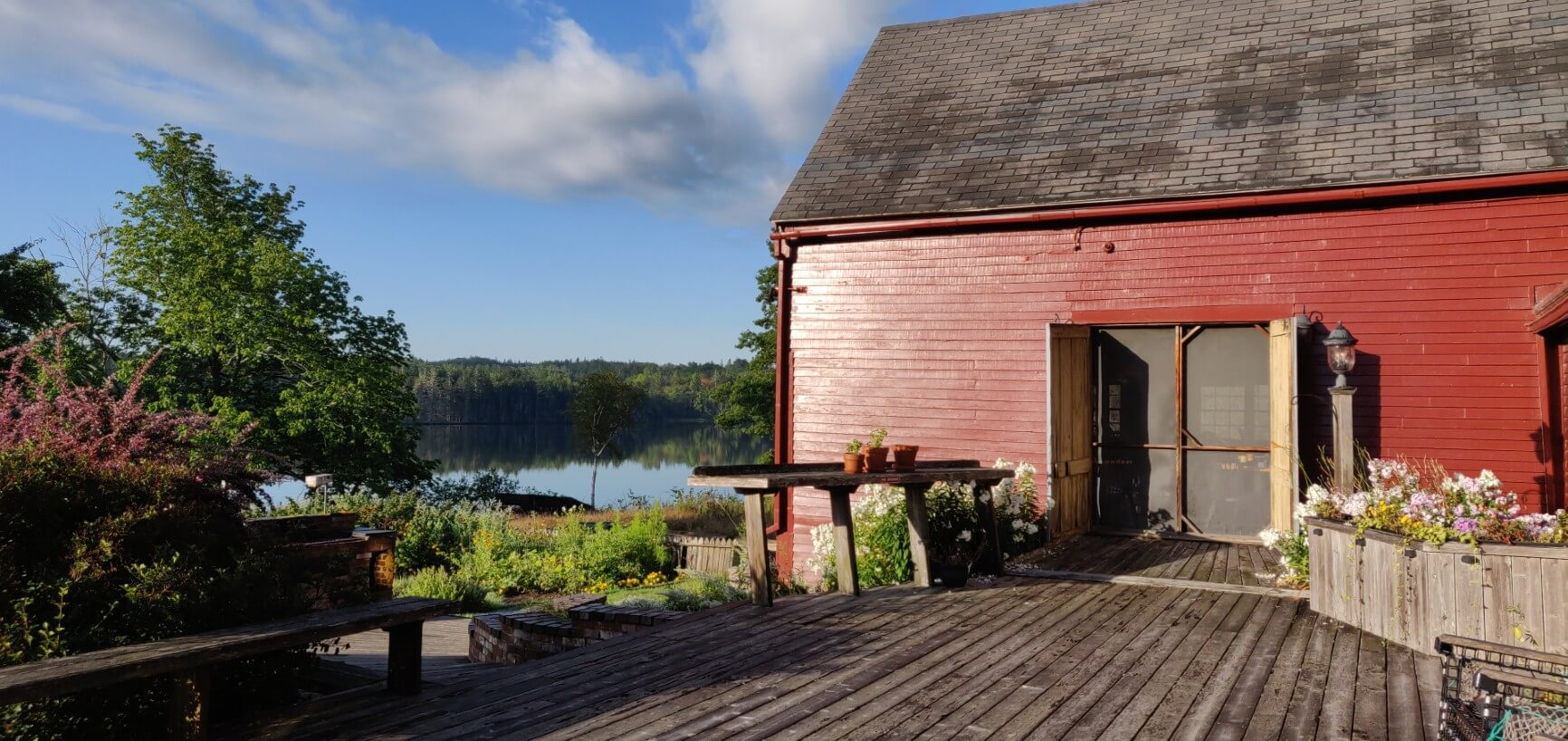 Red barn with doors open, deck, expansive gardens with view of water and trees beyond