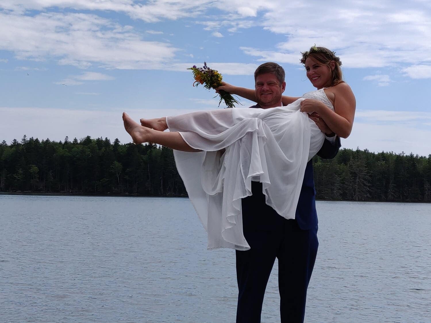 A groom carrying a bride with a bouquet with water, trees  and a blue sky in background