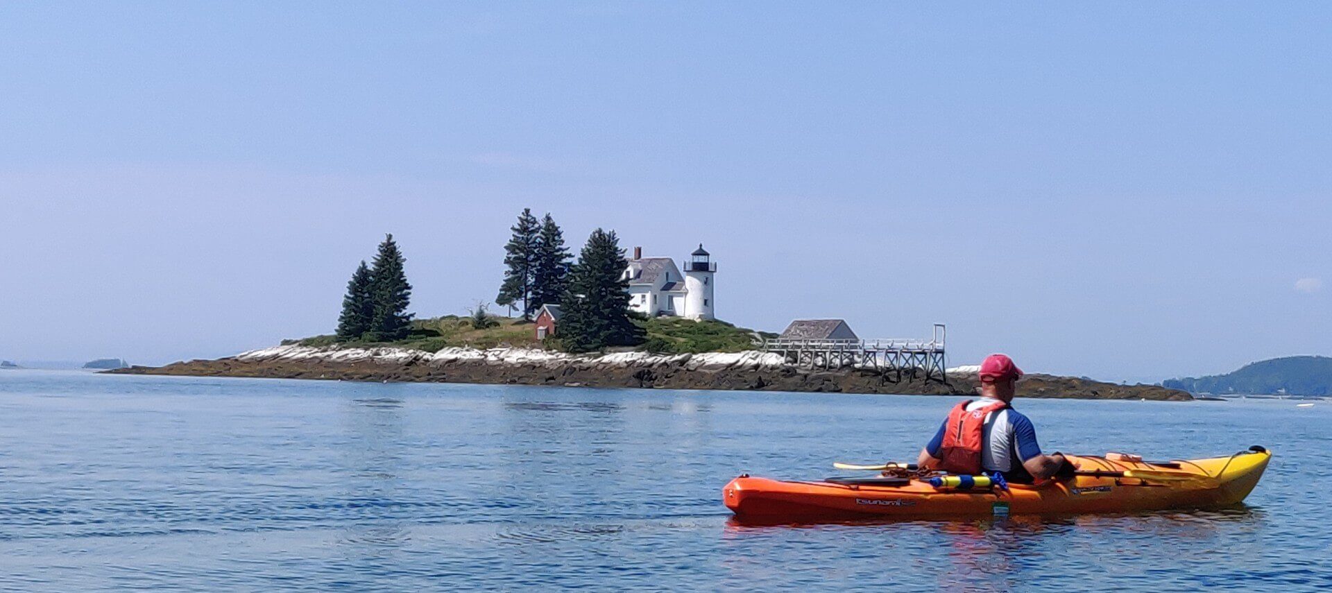A water view with a man in an orange kayak, and an island beyond that has trees, a house, and a lighthouse
