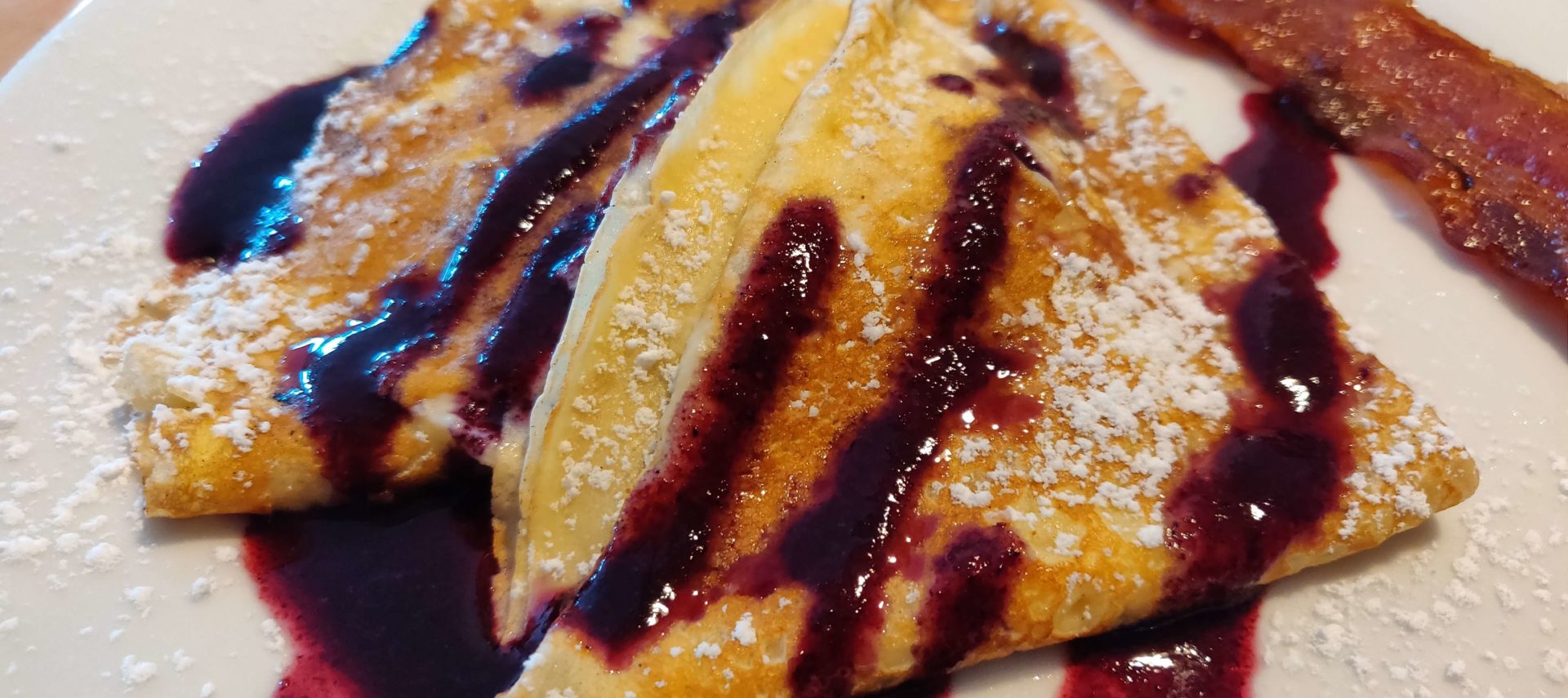 Crepes on a white plate with blueberry coulis and bacon