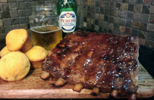 Slab of barbeque ribs on wooden board with cornbread muffins and bottle of Peroni beer behind