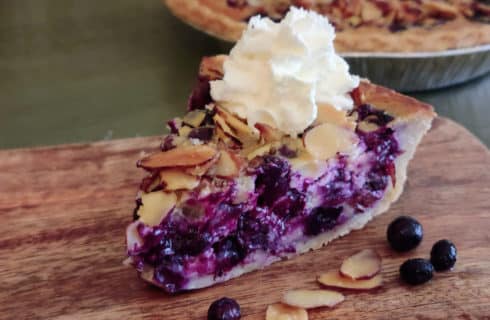 Slice of blueberry pie with almond slivers and whipped topping on wooded board