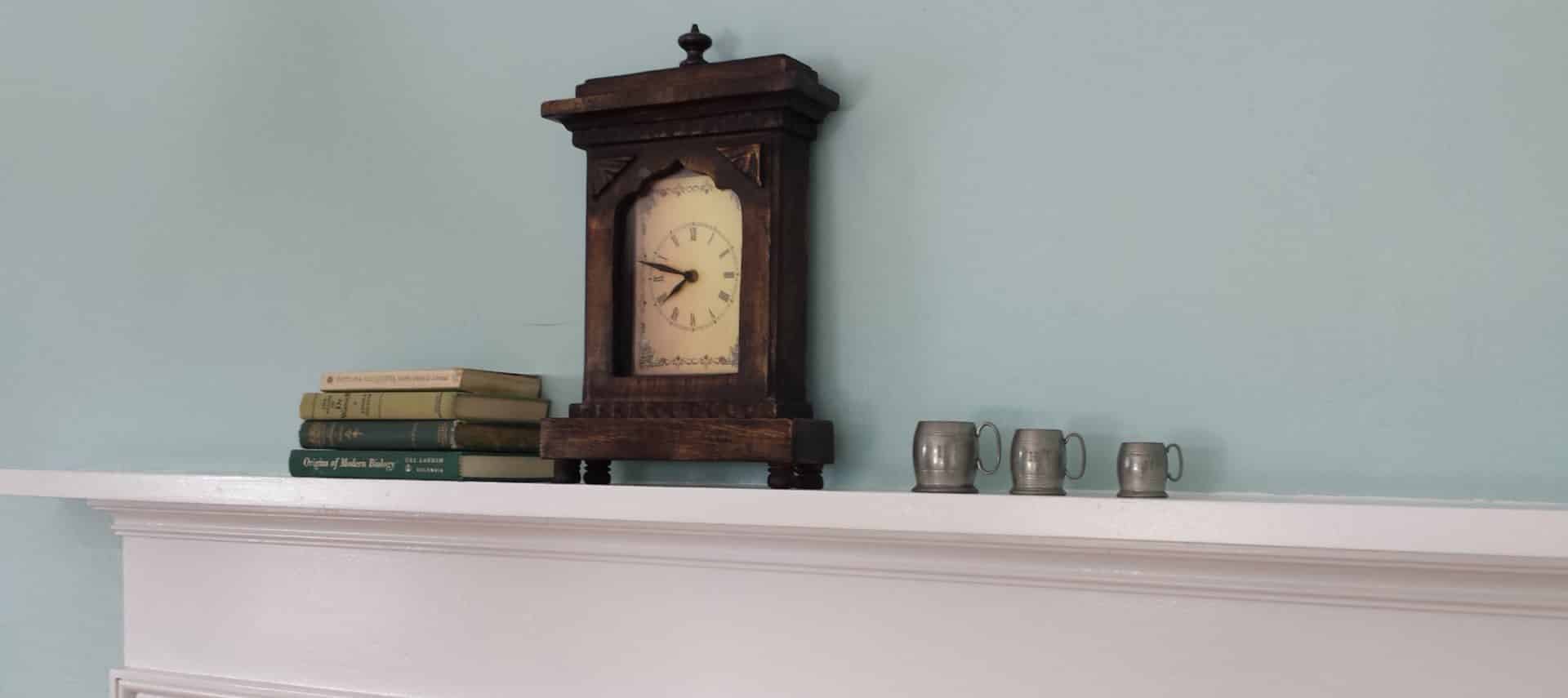 Close up view of old wooden mantel clock, books, and sliver cups on a white mantel with blue wall in the background