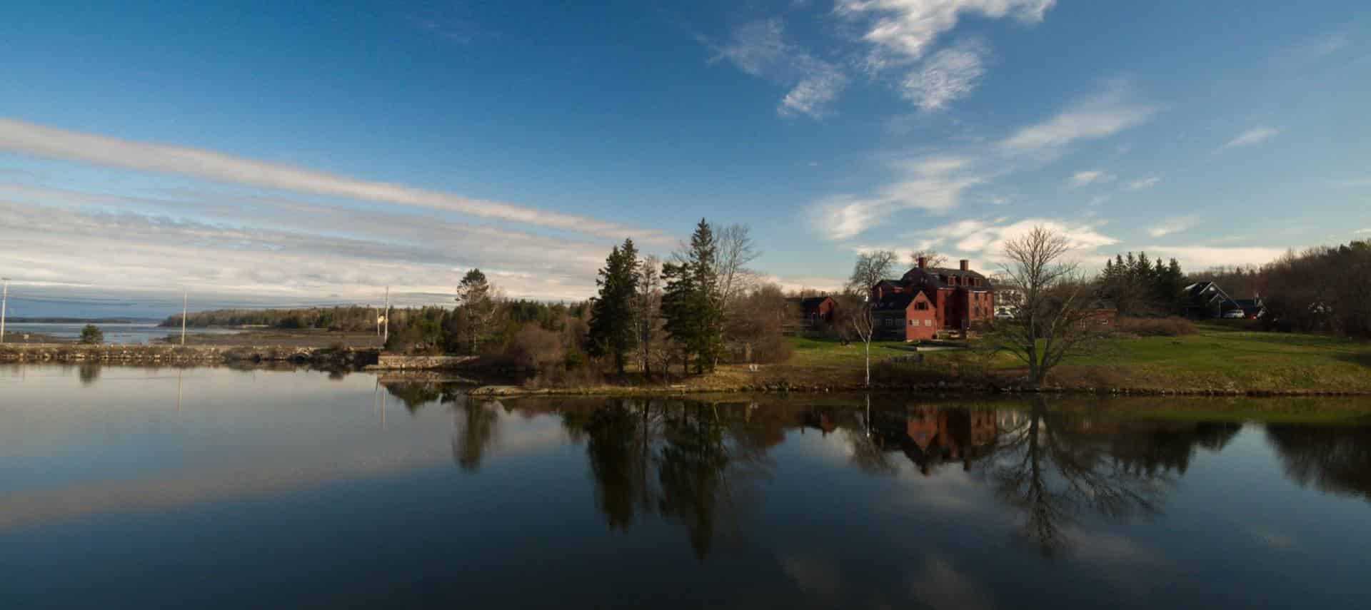 Exterior view of property surrounded by green grass and trees next to a large body of water with white clouds in the sky