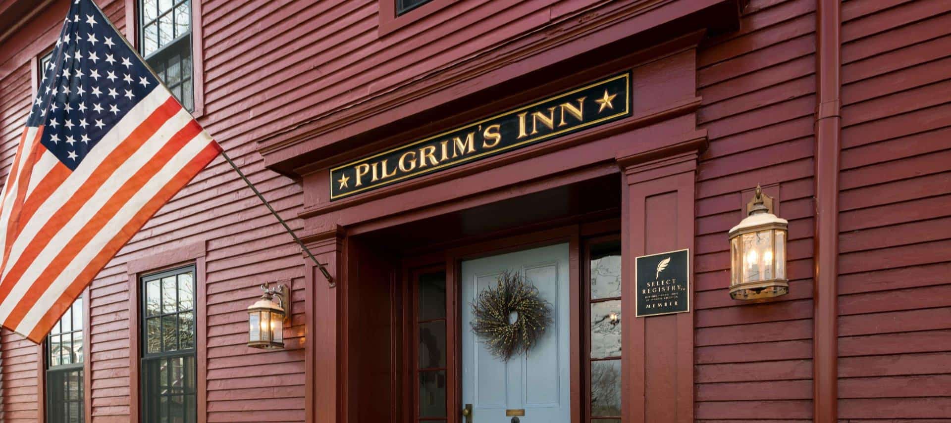 Close up view of the property painted dark red with a white front door, American flag, lantern sconces on each side of the entry, and large sign showing Pilgrim's Inn