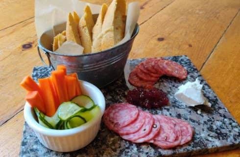 Carrot sticks and cucumber slices in white bowl, sliced bread in tin pail, sliced meat, soft cheese, and jelly on marble board